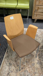 A modern chair features a light wood back, brown seat cushion, and metal legs. It has armrests and is set against a patterned rug and green stools.
