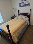 Cozy bedroom featuring a wooden bed with a yellow and gray comforter, nightstand with a lamp, and wall art above the bed. Plush carpet flooring.