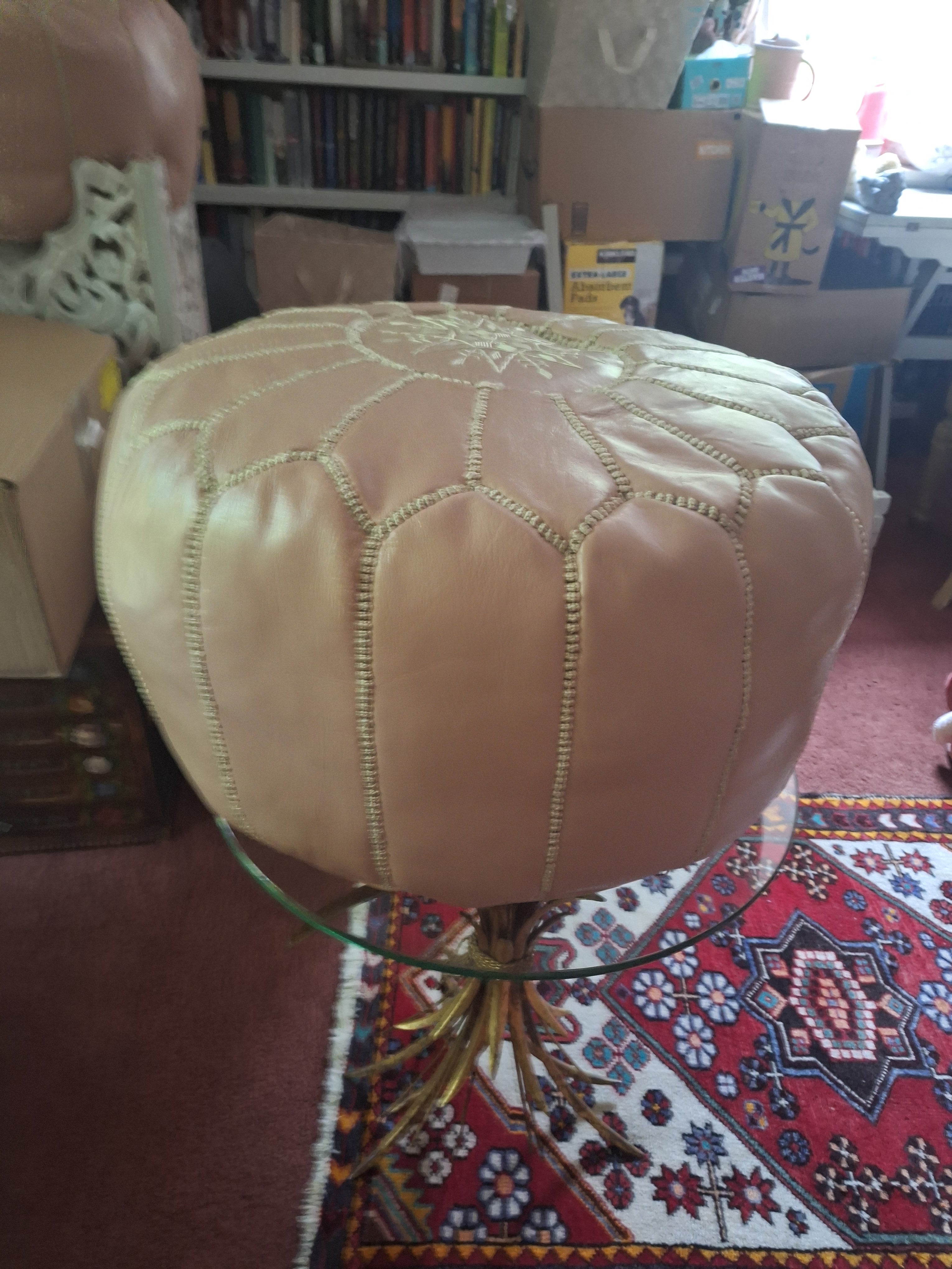 A round, beige leather pouf with decorative stitching is placed atop a glass table, surrounded by bookshelves and boxes in a cozy room.