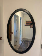 An oval mirror with a black frame reflects a well-lit hallway and a patterned rug, showcasing a bright, modern interior design.