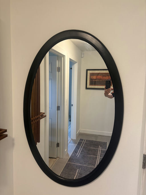 An oval mirror with a black frame reflects a well-lit hallway and a patterned rug, showcasing a bright, modern interior design.