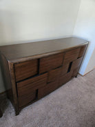 A modern wooden dresser with a sleek design featuring multiple geometric-shaped drawer fronts, sitting on a carpeted floor against a light wall.