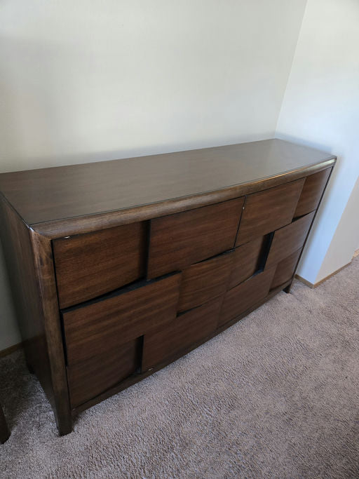 A modern wooden dresser with a sleek design featuring multiple geometric-shaped drawer fronts, sitting on a carpeted floor against a light wall.