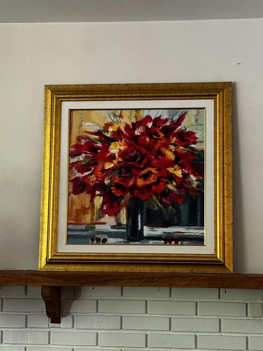 A vibrant painting of a red flower bouquet in a dark vase, framed in gold, hangs on a wall above a wooden shelf. The background features soft hues.