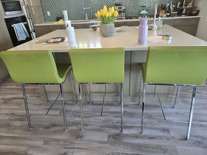 A modern kitchen scene featuring a sleek island with three green chairs, a vase of yellow tulips, and various kitchen items on the counter.