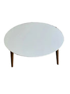 A modern round coffee table with a white top and wooden legs, placed on beige carpet, with a staircase visible in the background.