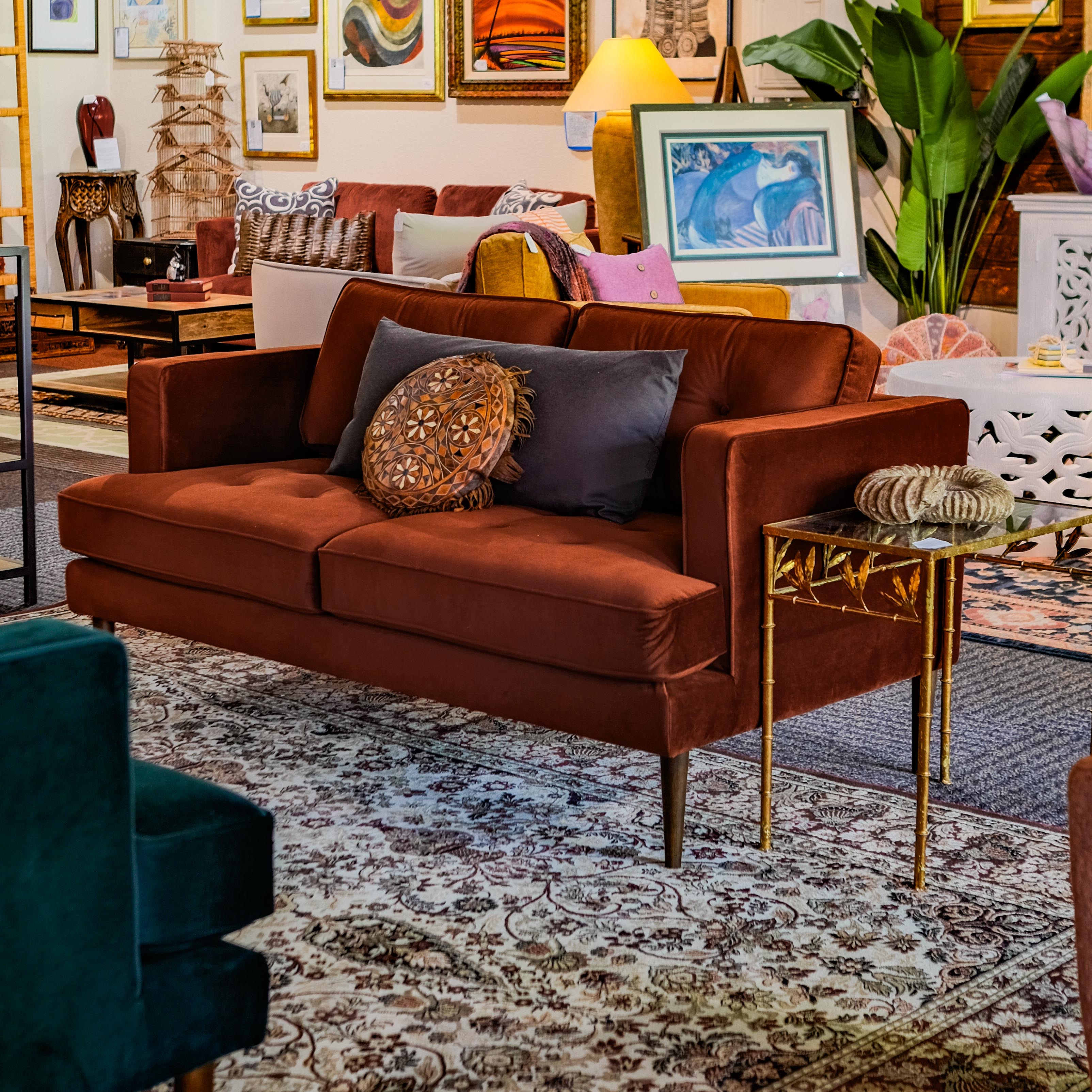A cozy, rust-colored velvet loveseat adorned with decorative cushions in an eclectic living room setup. The space is filled with a mix of contemporary and vintage furniture, artwork, and vibrant decor items, creating a warm and inviting atmosphere.