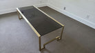 A modern coffee table with a sleek black glass top and gold metal frame sits on a neutral, textured carpet in a sparsely furnished room.