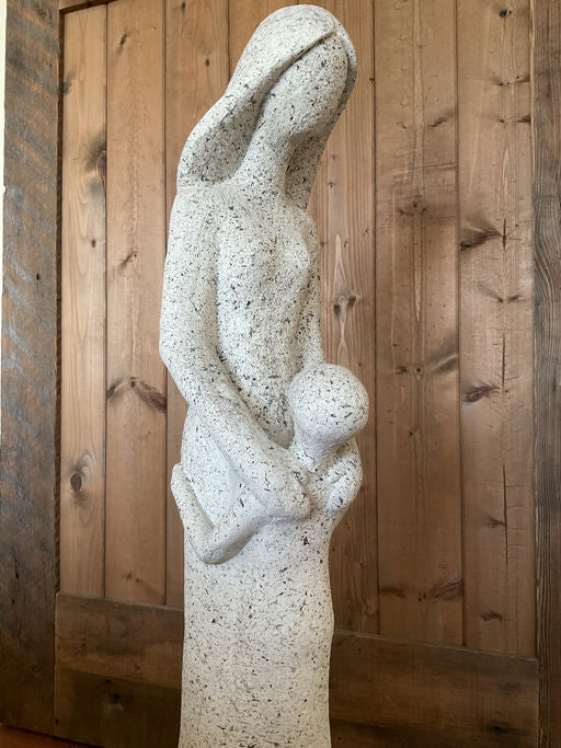A stylized stone sculpture of a woman holding a child close, conveying themes of nurturing and motherhood, set against a wooden background.