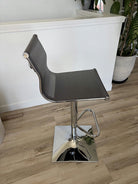 A modern bar stool with a sleek mesh seat and chrome base, positioned in a bright room featuring plants and a white wall.