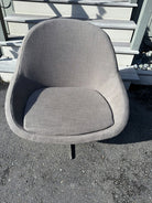 A gray, upholstered chair with a rounded back and cushioned seat sits on a gravel surface, in front of wooden steps and a pale wall.