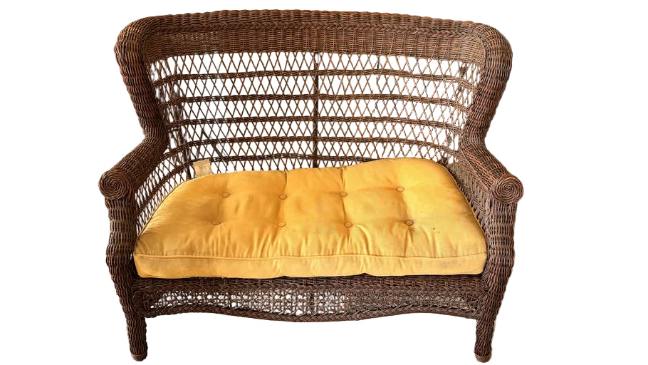 A wicker loveseat with a bright yellow cushion sits on carpet, near a dining table. A red backpack and a green bag are visible in the background.