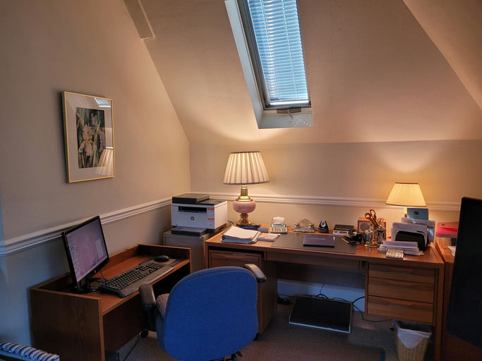 Home office with angled ceiling features a computer desk, printer, desk lamps, and decorative framed artwork, creating a cozy workspace.