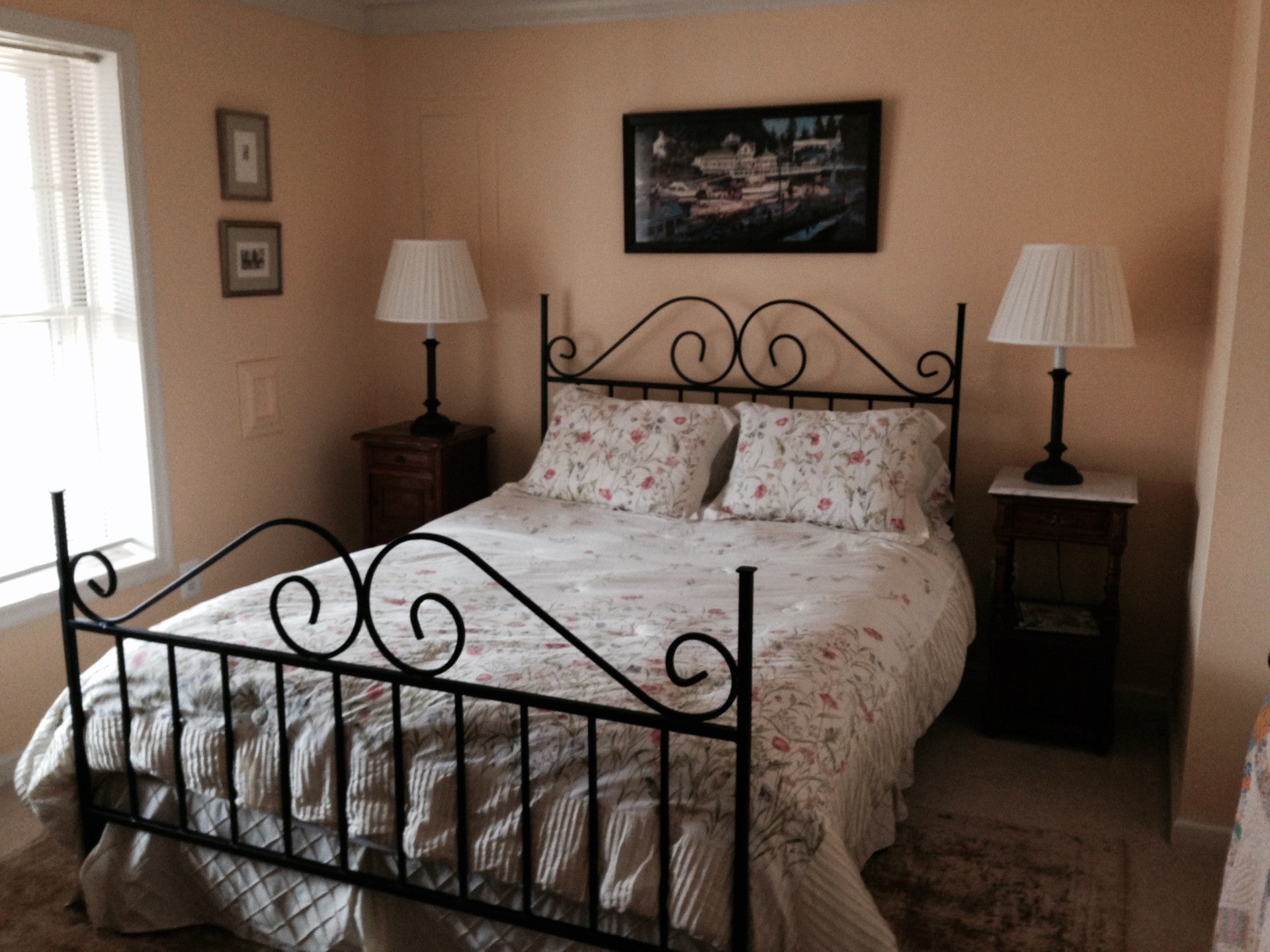 A cozy bedroom with a black iron bed, floral bedding, two bedside lamps, and a painting on the wall, illuminated by natural light from a window.