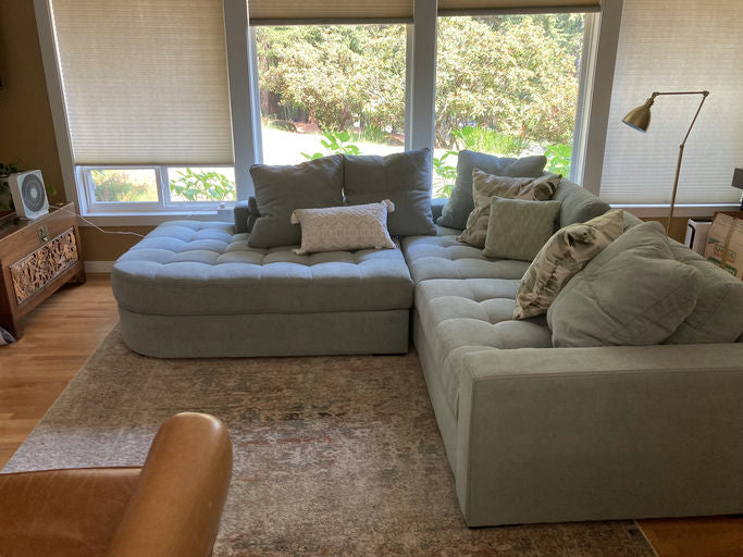 A cozy light blue sectional sofa with multiple pillows sits in a bright room, alongside a wooden cabinet and a fan near large windows.