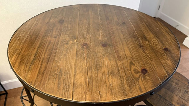 A round wooden table with a smooth, polished surface, featuring a rich grain pattern and a dark finish. Minimalist metal legs are visible.