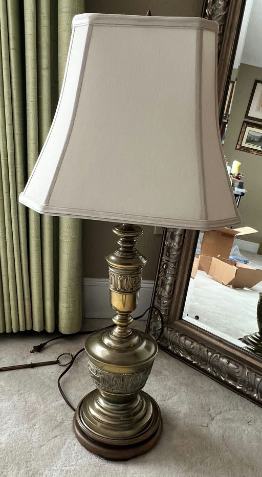 A vintage brass table lamp with a white, square shade stands beside a large ornate mirror, against a backdrop of green curtains and a light carpet.