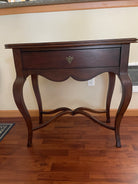 A wooden side table with curved legs and a single drawer, featuring a decorative handle, set against a neutral wall and wooden floor.