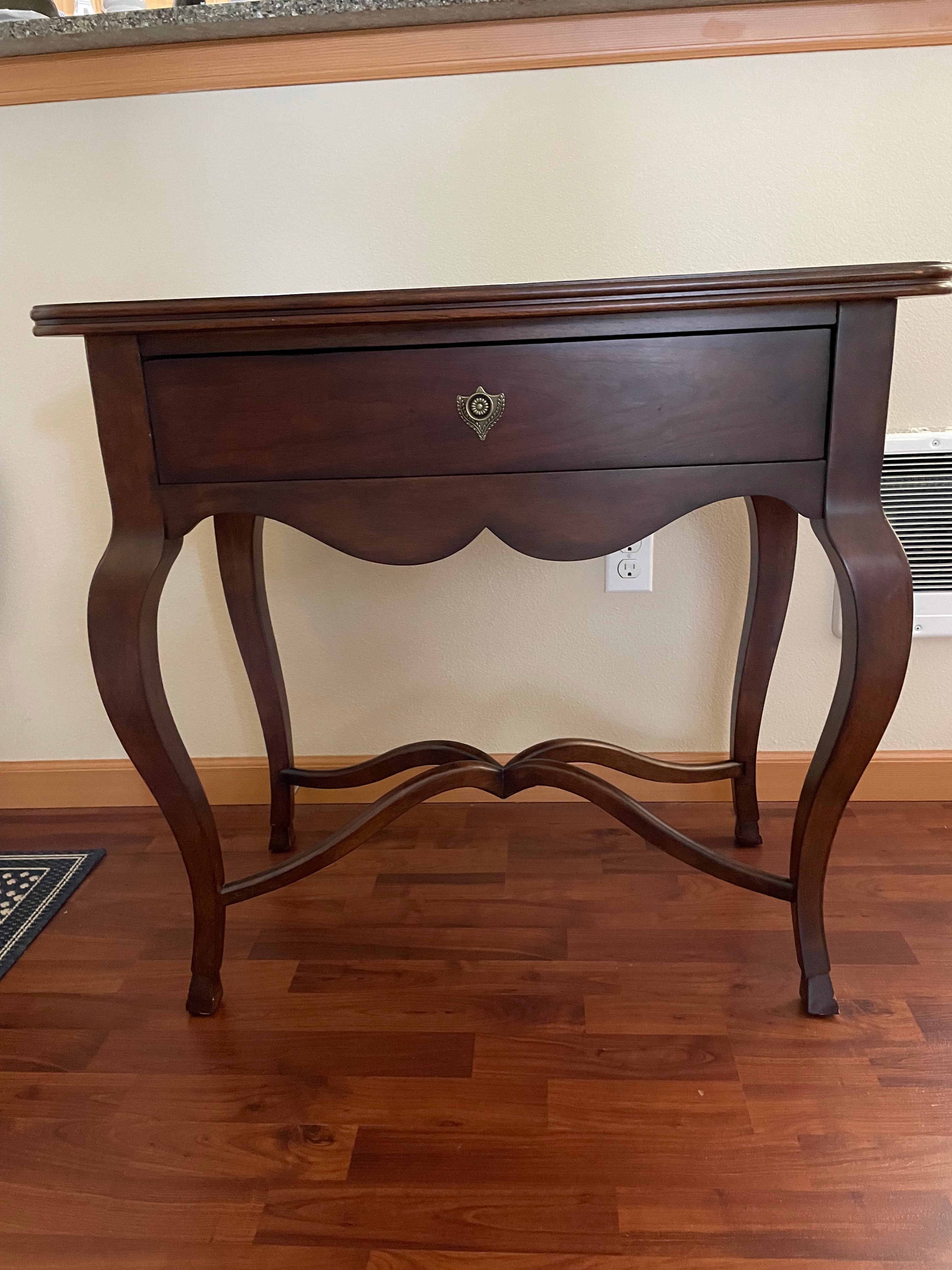 A wooden side table with curved legs and a single drawer, featuring a decorative handle, set against a neutral wall and wooden floor.
