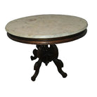 A round marble-top table with an ornate wooden base stands in a corner, next to a wall outlet, on a patterned wooden floor.