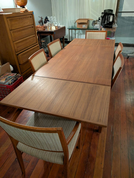 A wooden dining table with four upholstered chairs sits in a cozy room. Background elements include a dresser, glassware, and unpacked boxes.