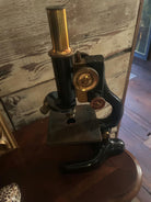 A vintage microscope with a brass and black finish sits on a wooden table, highlighting its intricate design against a rustic background.