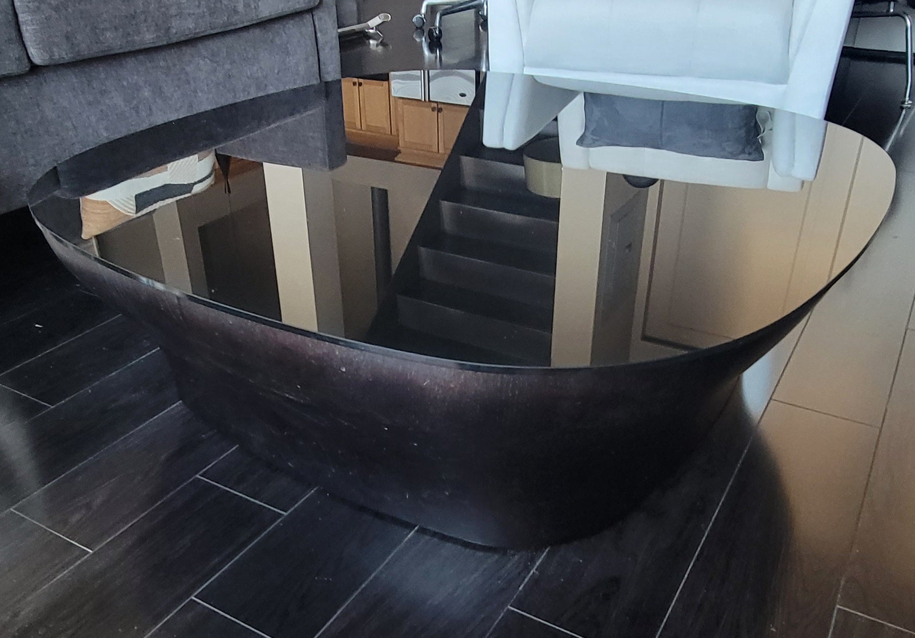 A modern, oval coffee table with a dark wooden base and reflective glass top is shown in a living room setting with a couch and staircase.
