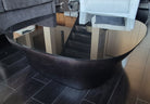 A modern, oval coffee table with a dark wooden base and reflective glass top is shown in a living room setting with a couch and staircase.