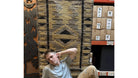 A young man poses against a patterned rug in a store, surrounded by boxes. The background features rustic decor and natural elements.