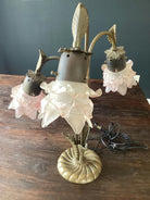 A decorative lamp with three flower-shaped glass shades in soft pink, featuring a brass base and leafy accents, placed on a wooden surface.