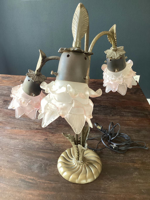 A decorative lamp with three flower-shaped glass shades in soft pink, featuring a brass base and leafy accents, placed on a wooden surface.