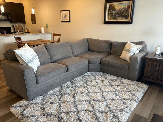 A cozy living room featuring a grey sectional sofa with decorative pillows, a plush rug, a wooden side table, and a dining area in the background.
