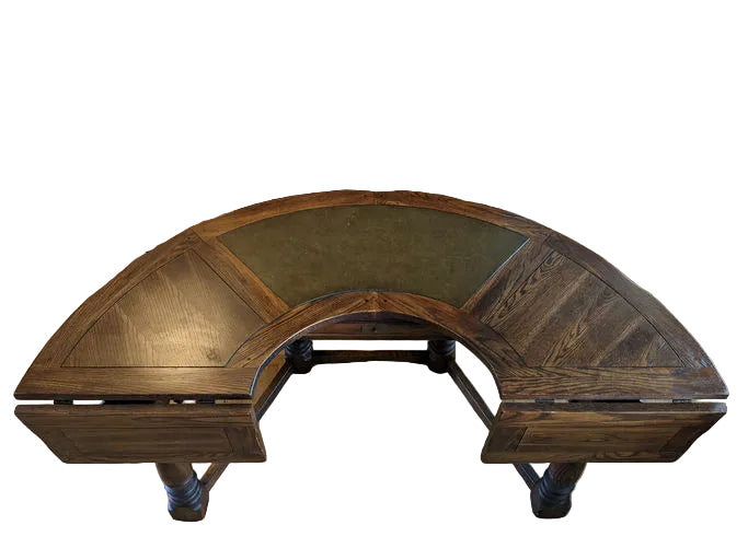 A curved wooden desk with a green leather inlay, featuring intricate detailing and storage drawers, set on a hardwood floor against a light wall.