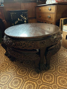 A carved wooden round table sits in a room with vintage decor, featuring intricate designs and a glossy surface, with patterned flooring visible.
