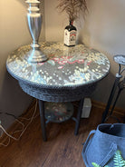 A round table with a floral design top, a silver lamp, a small bottle labeled “Pathfinder,” and a felt bag, situated beside a wooden floor.