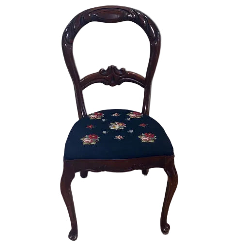A vintage wooden chair with a curved backrest and floral-embroidered navy seat, placed on a brown mat against a plain wall.