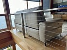 A modern living space featuring two light gray sofas on a wooden platform, separated by a stainless steel railing, with a view of the water outside.