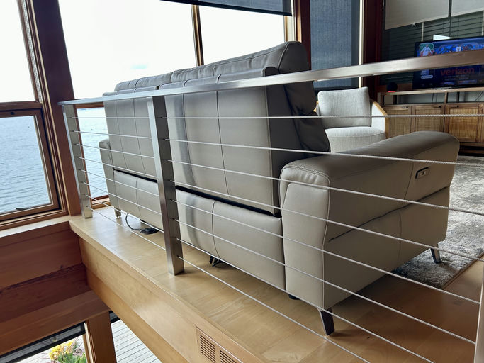 A modern living space featuring two light gray sofas on a wooden platform, separated by a stainless steel railing, with a view of the water outside.