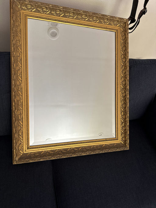 A vintage mirror with an ornate gold frame rests against a dark blue fabric sofa, reflecting light in a cozy indoor setting.