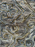 Close-up of a patterned rug featuring intricate swirls and geometric shapes in muted colors of blue, green, and brown. Soft texture visible.