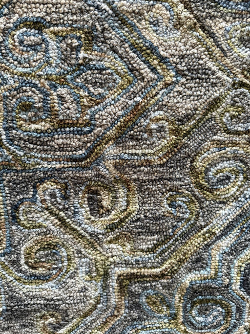 Close-up of a patterned rug featuring intricate swirls and geometric shapes in muted colors of blue, green, and brown. Soft texture visible.