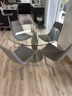 A modern dining area features a glass round table surrounded by four sleek gray chairs, set on light wooden flooring, creating an airy, elegant space.