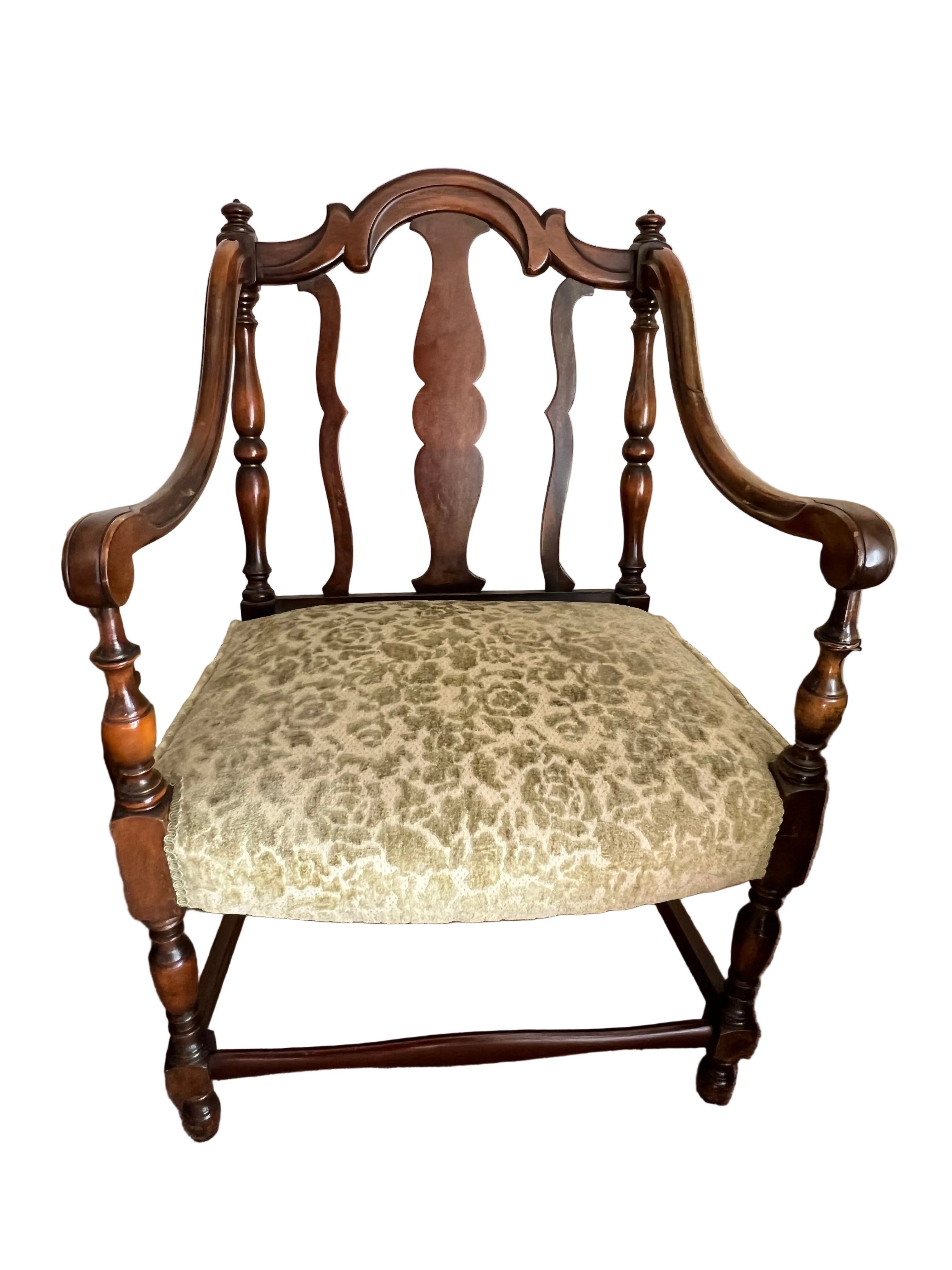 A vintage chair with a wooden frame, featuring an ornate backrest and green patterned upholstery. It rests on a carpeted floor beside a table.