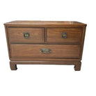 A wooden chest of drawers with two small and one larger drawer, featuring metal handles, placed on a concrete surface. A garden setting is in the background.