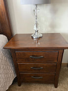 A wooden nightstand with three drawers, featuring black handles. A crystal lamp with a gray shade is placed on top, beside a bed with a quilted cover.