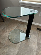A modern, two-tier glass side table with a black cylindrical base. The top surface is oval-shaped, resting beside a window on a tiled floor.