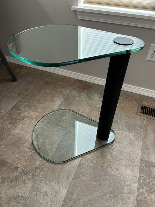 A modern, two-tier glass side table with a black cylindrical base. The top surface is oval-shaped, resting beside a window on a tiled floor.