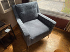 A cozy gray armchair sits near a window, with an air conditioning unit above and a wooden side table beside it, casting soft shadows on the floor.