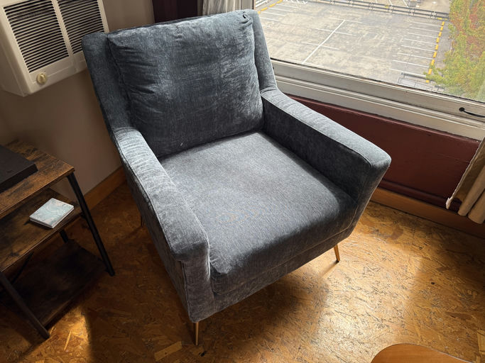 A cozy gray armchair sits near a window, with an air conditioning unit above and a wooden side table beside it, casting soft shadows on the floor.