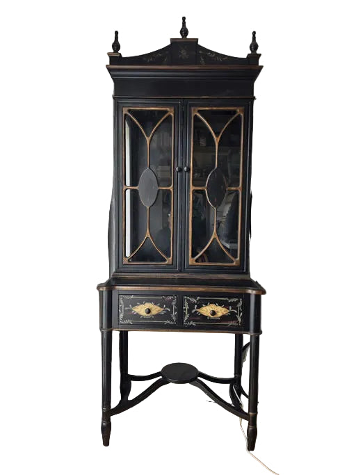 A vintage black cabinet with glass doors and decorative details stands in a well-lit room, featuring a lower drawer with floral designs.